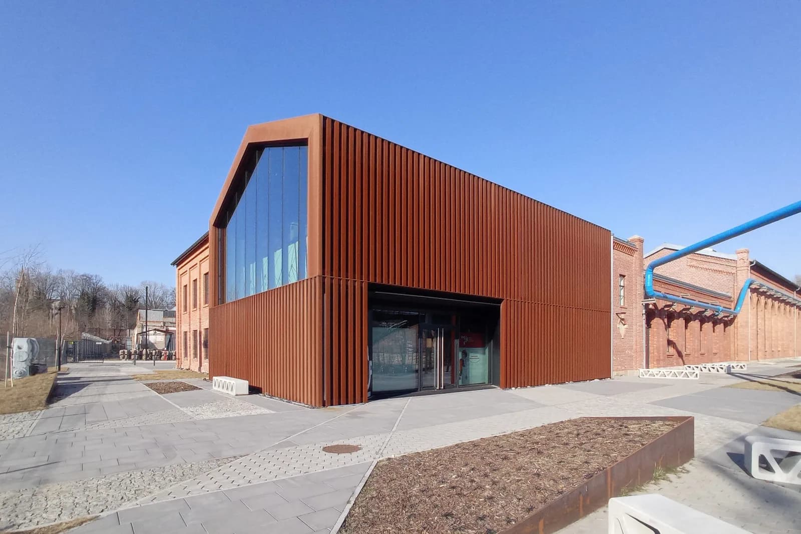 A modern building with a rusted metal facade and a large glass window on the left, next to an older brick building with a bright blue pipe running along its side, under a clear blue sky.